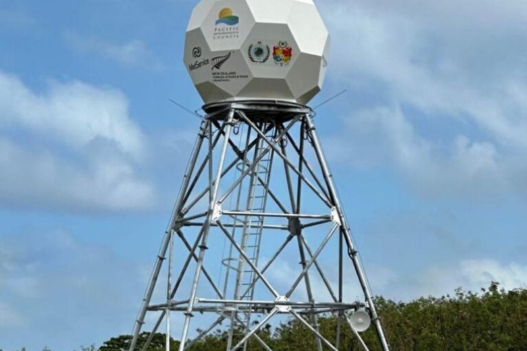 A geodesic dome-shaped weather radar tower stands on a metal framework against a background of blue sky and clouds. Logos are visible on the dome's surface.