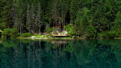 A serene lake reflecting a dense forest with a wooden structure and people at the water's edge, surrounded by tall evergreen trees.