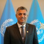 A person in a suit stands in front of two blue flags with the United Nations emblem.