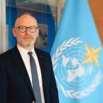 A man in a suit and tie stands next to a flag with the United Nations emblem in an office setting.